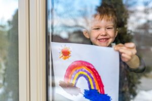 Young Boy sticking his rainbow drawing on home window during the Covid-19 crisis